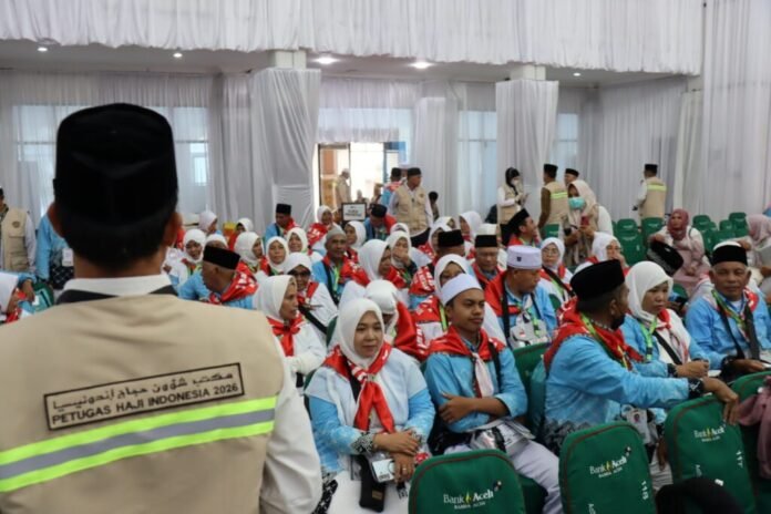 An orderly airport arrival hall in Indonesia where Hajj pilgrims wearing white traditional garments and identification lanyards walk through a clearly marked corridor while uniformed health officers conduct medical screenings, with families wearing surgical masks waiting patiently behind designated barriers under bright overhead lighting and natural daylight from large terminal windows.