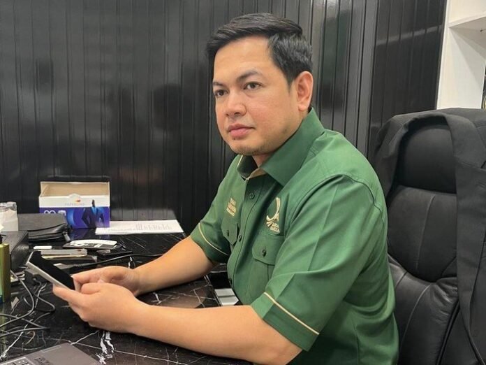 Tommy Kurniawan speaking at a public event, wearing formal attire, with the MBG program logo in the background, realistic style, clear daylight setting, professional news conference atmosphere